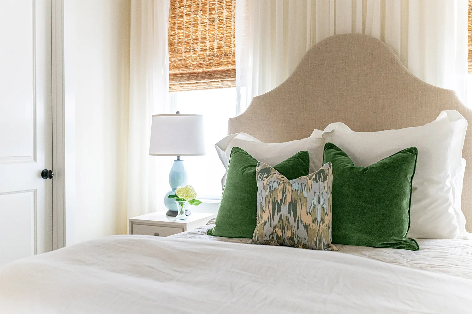 Bed with an arched oatmeal linen headboard, white bedding and green velvet pillows beside a sunlit window