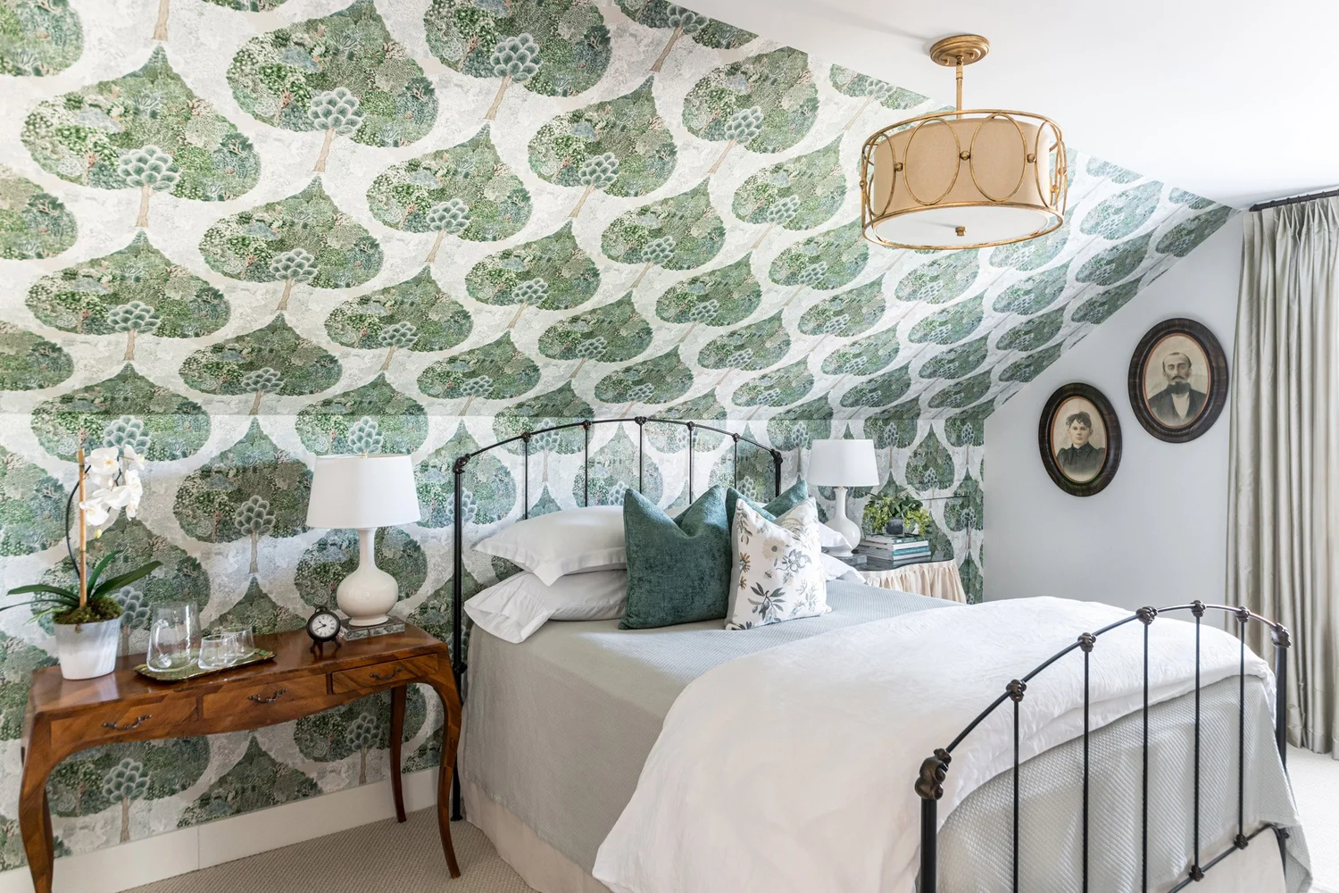 Black iron bed with white linens under a sloped ceiling papered in a green leafy tree pattern