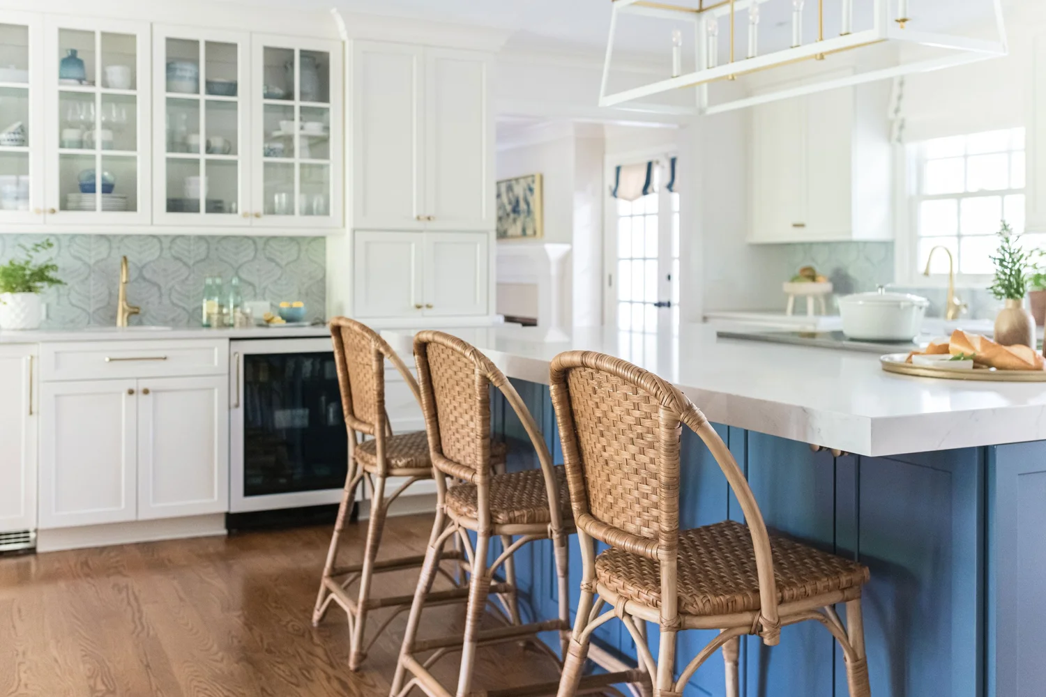 White kitchen with a blue island, marble waterfall counter and three rattan counter stools on oak floors