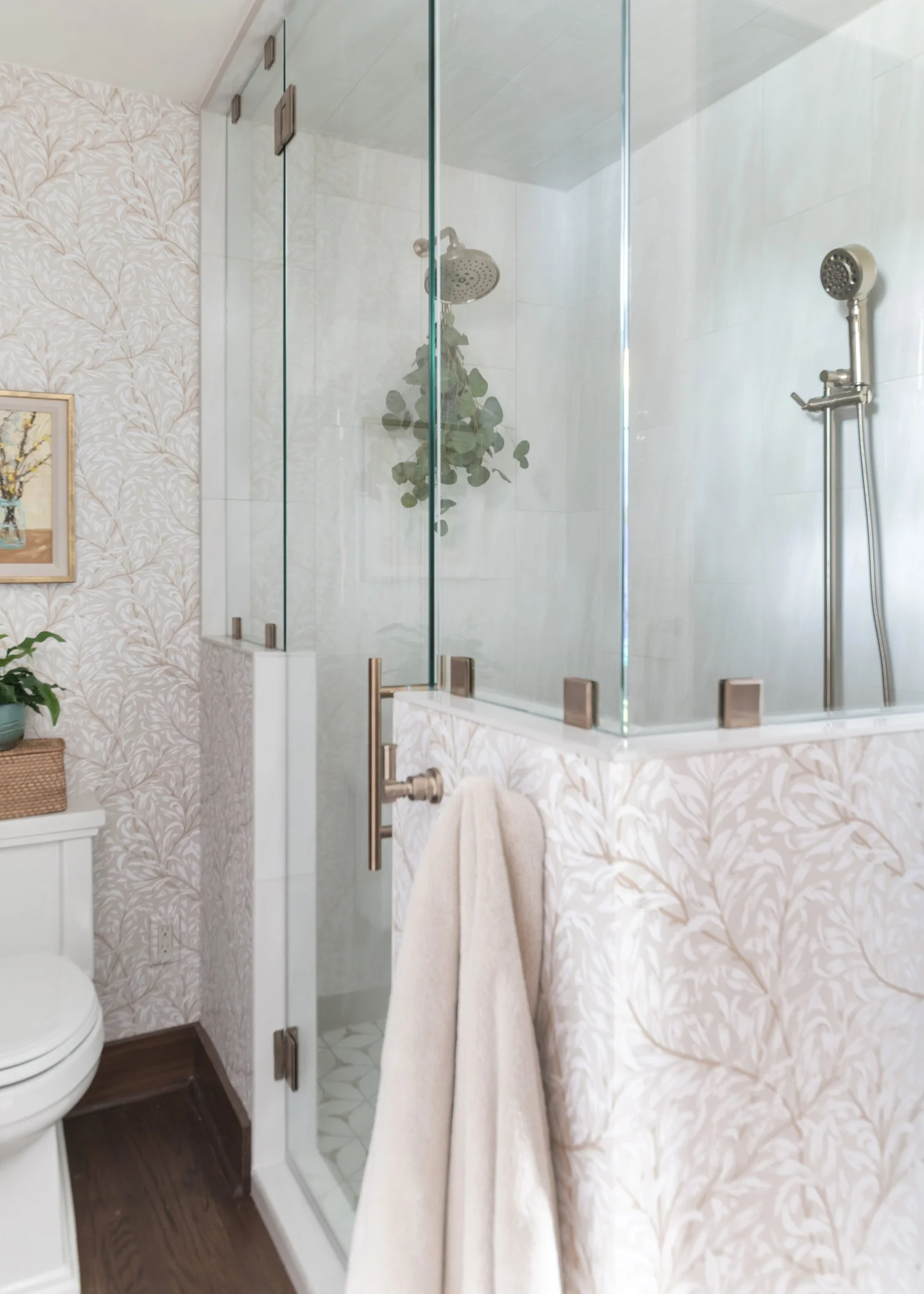 Glass-enclosed shower with large-format marble-look tile, beside blush leaf wallpaper and a white toilet