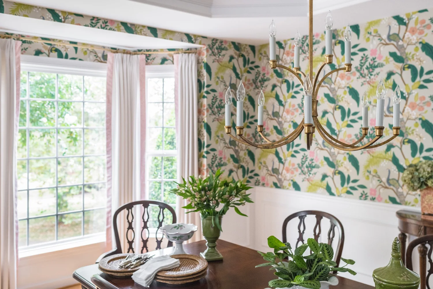 Dining room in bold floral-and-toucan wallpaper above white wainscot, with a mahogany table and gilt chandelier