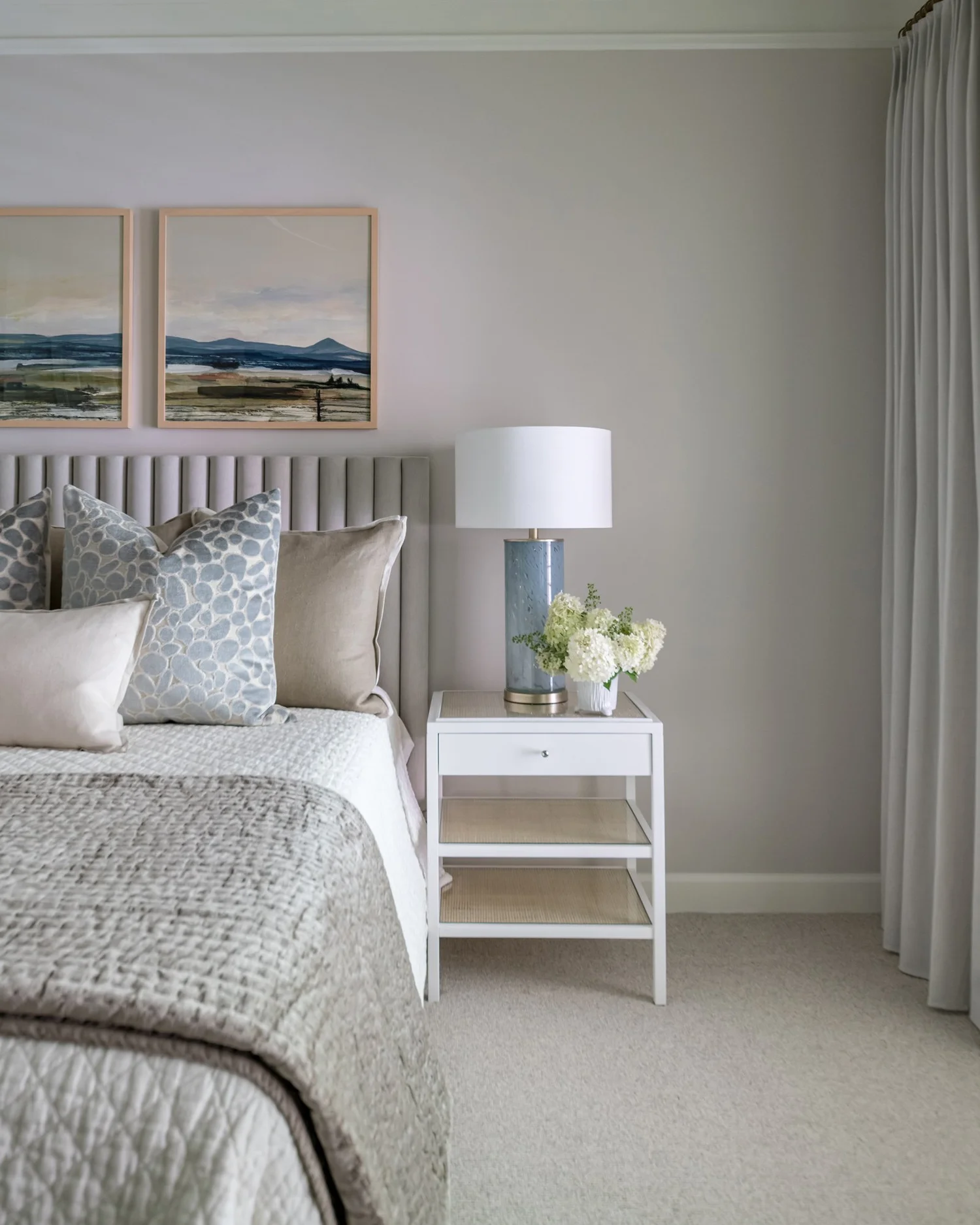 Bed with a channel-tufted grey headboard and quilted coverlet next to a white cane nightstand on pale carpet