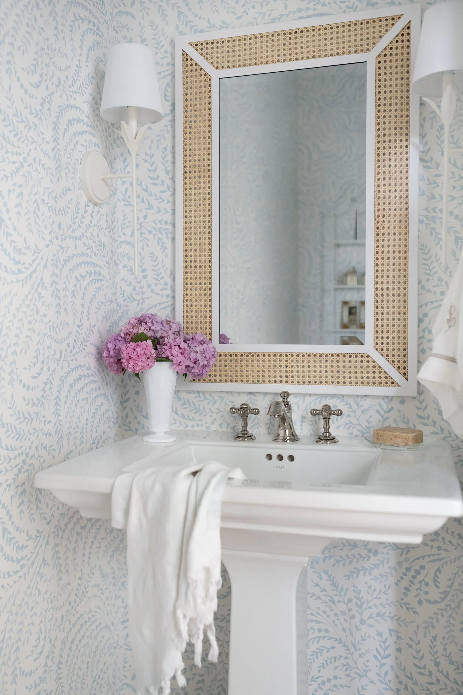 White pedestal sink against pale blue botanical wallpaper, under a cane-framed mirror with a linen towel draped over